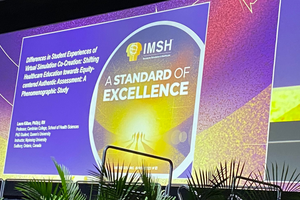 image of an empty conference stage with a presentation slide from the Society for Simulation in Healthcare projected. There are decorative palm plants along the bottom of the stafe.